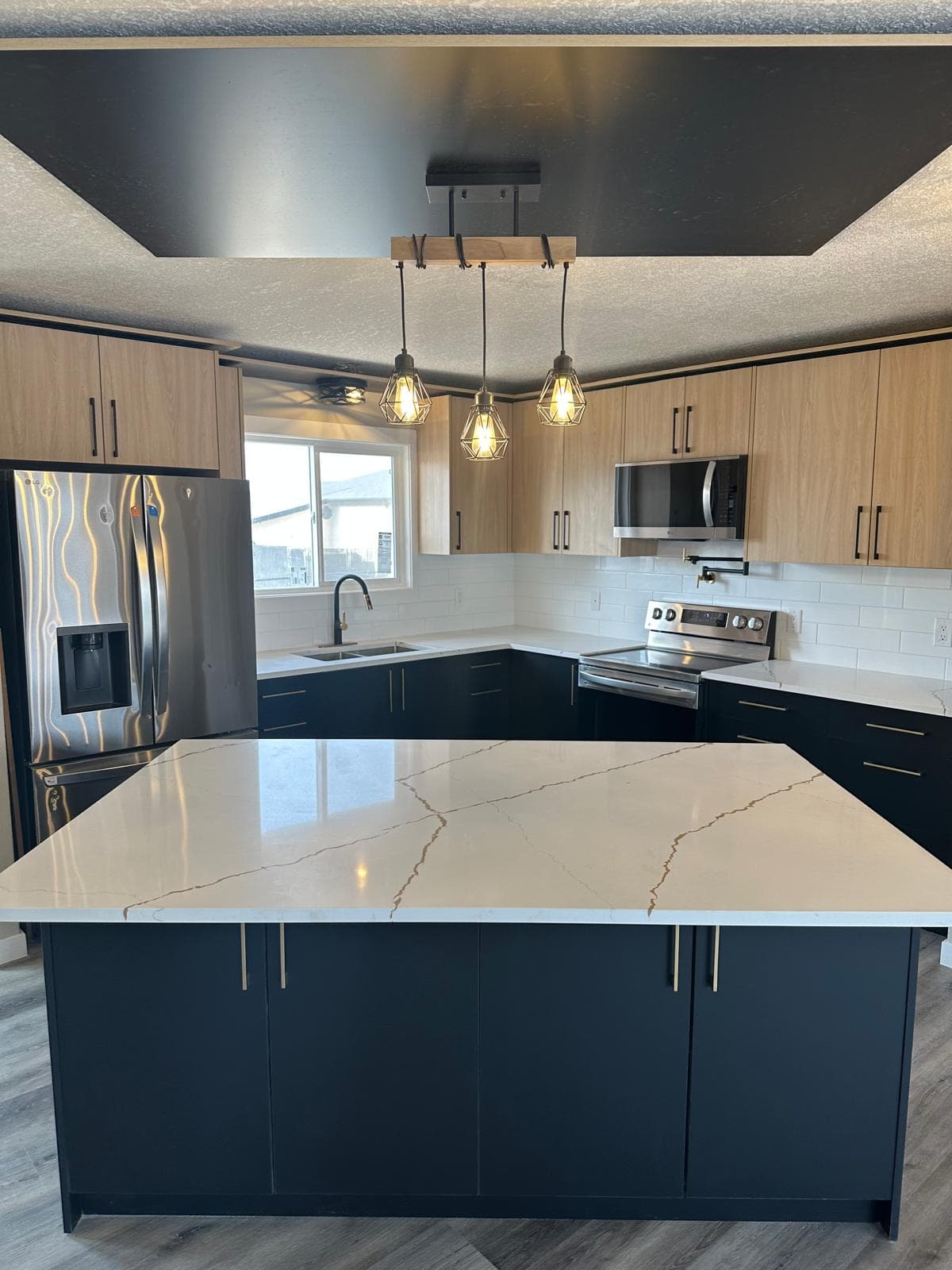 JFK Surfaces Ponoka kitchen renovation — black waterfall quartz island with stainless LG French-door fridge, two-tone cabinets, and luxury vinyl plank flooring