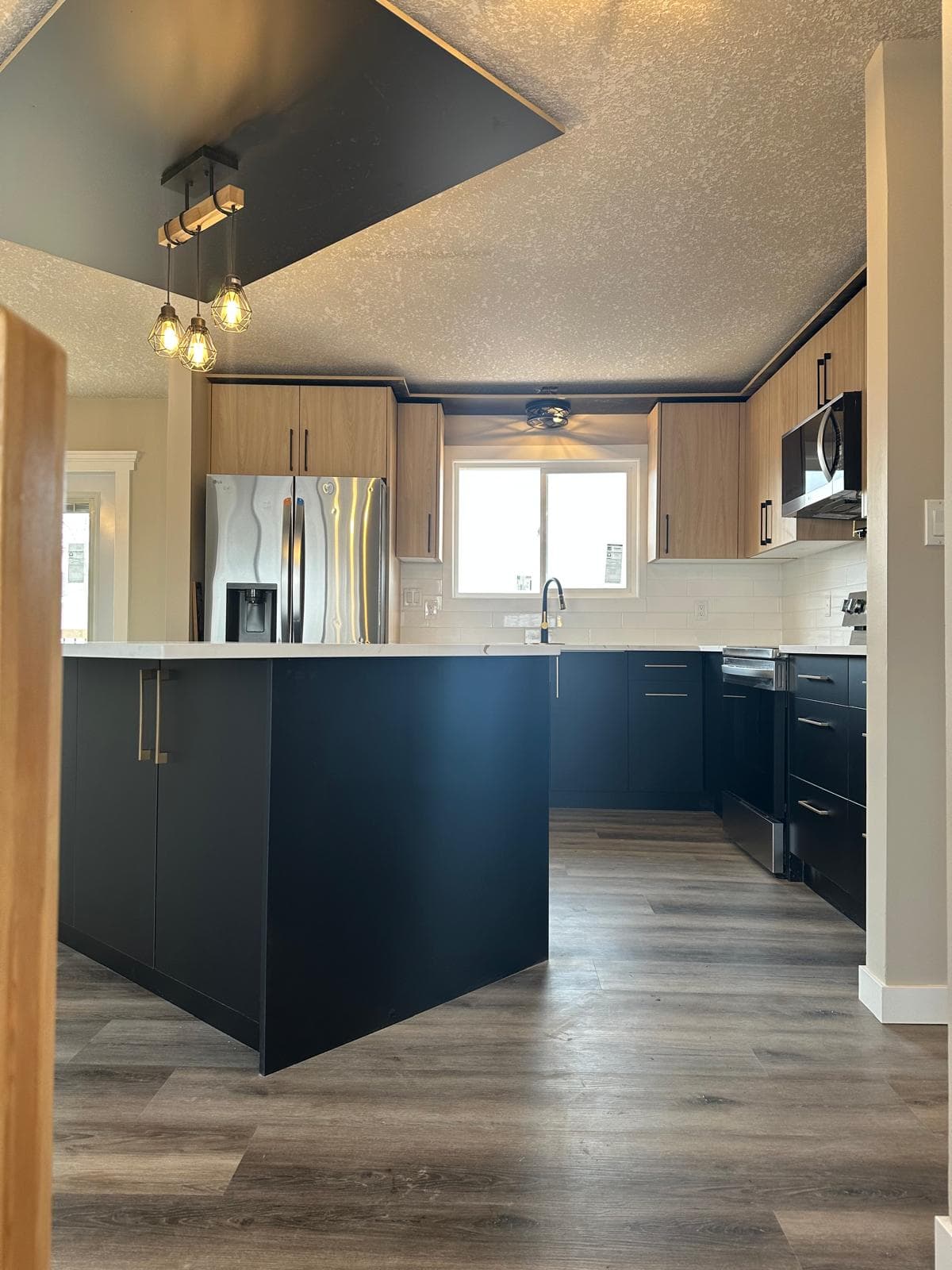 Ponoka kitchen renovation by JFK Surfaces — two-tone black and natural-wood cabinets, calacatta quartz waterfall island, custom black ceiling cove with cage pendants, brass pot filler, white subway tile backsplash